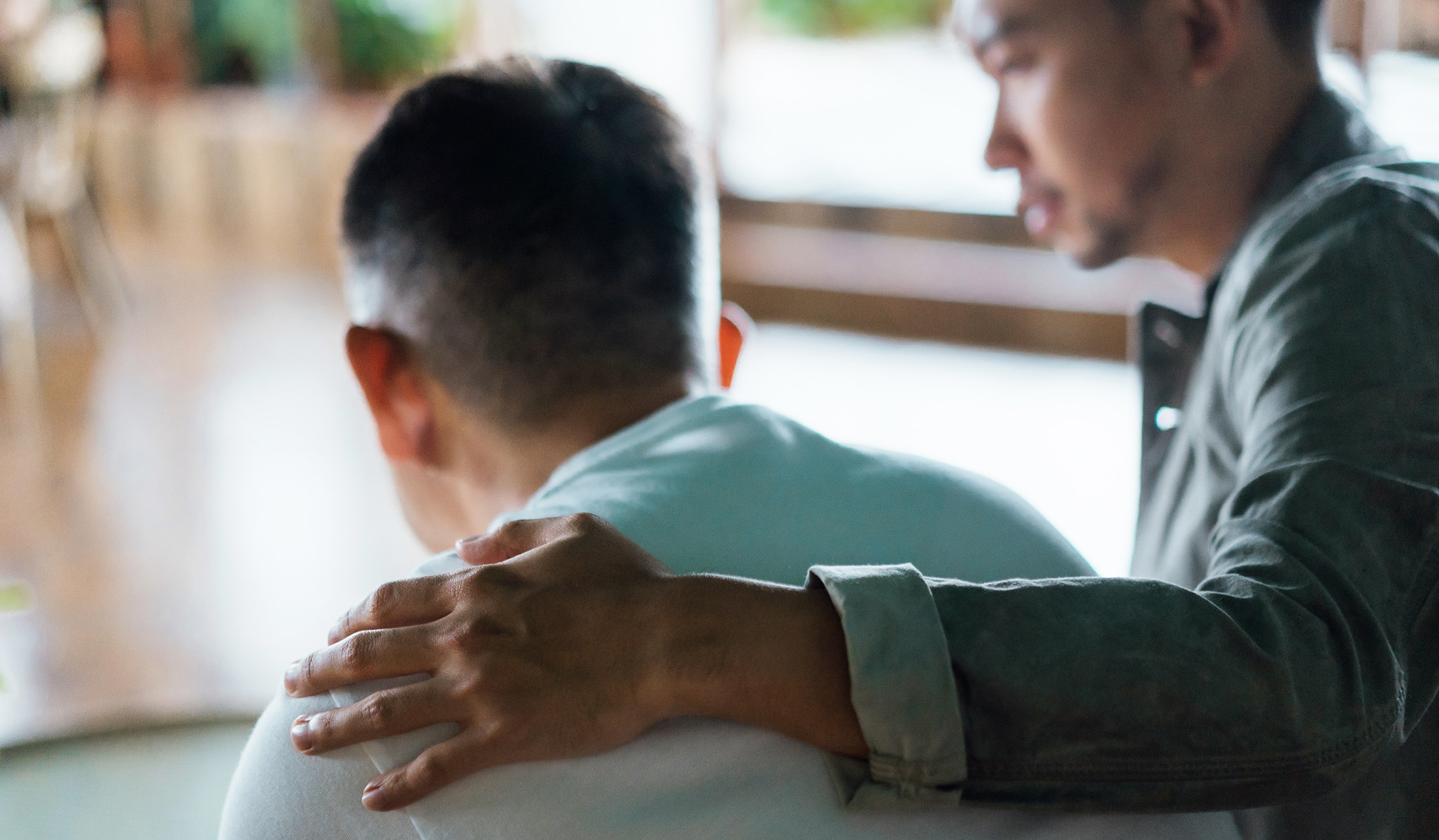 Rear view of son and elderly father sitting together at home. Son caring for his father, putting hand on his shoulder, comforting and consoling him. Family love, bonding, care and confidence