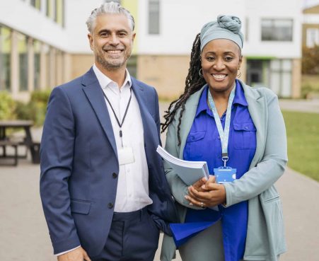 Portrait of cheerful teachers on secondary school campus Front view of British man and woman in early 50s standing side by side and smiling at camera, education building in background.